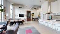 This single-floor communal space showcases a contemporary open-plan design with clean white walls, recessed lighting, and stylish pendant lights that illuminate the room evenly. The layout is thoughtfully divided into distinct functional zones while maintaining a seamless flow throughout. A comfortable lounge area anchored by a gray sectional sofa and a geometric patterned area rug with a bold pink accent sits alongside a dining arrangement with a rectangular table and matching chairs. A wall-mounted flat-screen television serves as a focal point in the lounge zone, flanked by abstract linear wall art. Toward the center of the space, a pool table provides a recreational focal point, complemented by additional colorful abstract artwork on the surrounding walls. On the opposite side, a sleek white bar counter with modern bar stools creates a casual social gathering spot. Light-toned tile flooring runs throughout, enhancing the bright, airy ambiance of the space. The combination of neutral tones with vibrant art pieces, varied seating arrangements, and multi-purpose zones makes this an inviting and versatile communal area well-suited for socializing and recreation.