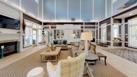 Located at 3960 Bell Rd in Nashville, TN, this single-floor community clubhouse presents an inviting and well-appointed gathering space designed with both comfort and functionality in mind. The entryway makes a strong first impression, featuring large glass-paneled doors framed by transom windows that flood the foyer with natural light. Soaring double-height ceilings clad in soft blue tones, accented by crisp white paneling and rich brown wall treatments, establish a refined yet approachable architectural character throughout the space. The grand lounge area is generously proportioned, offering multiple comfortable seating arrangements with neutral-toned sofas and accent chairs accented by teal and gold decorative pillows. A built-in shelving unit provides both display and storage, while a wall-mounted television and a classic white fireplace with decorative tile surround serve as focal points. Large windows on multiple walls ensure the interior remains bright and connected to the surrounding outdoor environment. The leasing or reception area is thoughtfully integrated near the entrance, framed by white columns and paneled partitions that provide definition without sacrificing the open feel. Large-format tile flooring with geometric inlay detailing anchors the space and transitions seamlessly between functional zones, creating a cohesive and polished overall aesthetic.