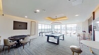 Located at Culebra Commons in San Antonio, Texas, this contemporary game and social lounge room is thoughtfully designed for recreation and relaxation. The space is anchored by a sleek, modern billiards table positioned beneath a striking circular chandelier that hangs from a gold-accented tray ceiling, adding a refined touch to the room's overall aesthetic. Plush, patterned carpeting in neutral tones runs throughout, complementing the clean white walls and contemporary décor.

A dedicated poker area features a round gaming table surrounded by comfortable upholstered chairs, accompanied by a large wall-mounted screen — ideal for both card games and media viewing. Nearby, a casual nook with two high-top tables, bar stools, and a built-in oversized Connect Four game set against a dark, textured accent wall creates an inviting space for social interaction.

Floor-to-ceiling windows line an expansive lounge area, flooding the space with natural light and offering sweeping views of the San Antonio skyline and surrounding community. Bistro-style tables and modern chairs are arranged to take full advantage of these panoramic vistas. Curated wall art and thoughtful lighting throughout tie together this well-appointed, versatile amenity space.