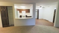 This single-floor residence is designed with an open and functional layout that maximizes the use of its living space. The kitchen is positioned behind a half-wall pass-through, creating a semi-open connection to the main living area. It features rich, dark wood cabinetry offering ample storage, white appliances including a refrigerator, microwave, and range, as well as light-toned countertops that brighten the space. The pass-through counter also doubles as a casual dining or prep surface, adding to the kitchen's practicality.

The living area extends beyond the kitchen wall, finished with warm carpeting that adds comfort underfoot. A ceiling fan is centrally mounted, providing both lighting and air circulation throughout the room. Hardwood-style flooring is visible in the transitional areas, offering a clean contrast to the carpet. Neutral wall tones in soft grey and white keep the atmosphere light and adaptable to a variety of interior styles.

A doorway visible from the main living area leads to additional rooms, with a glimpse of a closet suggesting well-integrated storage solutions. A baseboard heater along the far wall indicates a practical heating setup. Overall, the space presents a clean, straightforward, and functional residential environment.