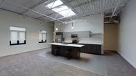 This single-floor space showcases a striking industrial-modern aesthetic, defined by its soaring ceiling height with exposed steel truss framework and a large skylight that floods the interior with abundant natural light. The open-concept layout creates a seamless, airy atmosphere throughout.

The kitchen is a central highlight of the space, featuring a large central island with a white countertop and built-in sink, complemented by dark wood cabinetry and bar-height stools for casual seating. A full suite of stainless steel appliances — including a refrigerator, stove, and microwave — is integrated into the upper and lower cabinetry, which carries a contemporary gray-toned finish. Pendant lighting hangs above the island, adding warmth and focused illumination.

The flooring transitions between polished concrete and wood-look tile, reinforcing the modern industrial character of the space. Track lighting along the exposed ceiling structure provides additional ambient and task lighting throughout. A window with a roller shade is visible on one wall, offering a view into an adjacent area. The neutral color palette of whites and grays throughout creates a clean, versatile environment suitable for a variety of uses.