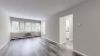 This single-floor unit offers three bedrooms, one bathroom, a kitchen, hallway, and closet within a thoughtfully arranged floor plan. The main living area is distinguished by light grey walls and durable hardwood-style plank flooring in a cool grey tone, with natural light streaming through wide, horizontally-oriented windows fitted with white blinds. A wall-mounted air conditioning unit and baseboard heating system serve the space. An arched doorway connects the living area to the kitchen, which features white shaker-style cabinetry, granite-look countertops, and matching hardwood-style flooring.

The spacious primary bedroom includes hardwood-style flooring, a built-in closet with bifold doors, and direct access to the hallway. The two additional bedrooms are carpeted and each receive natural light through windows with wooded outdoor views, with baseboard heaters beneath. The shared bathroom is finished in white tile, featuring a pedestal sink, full tub-shower combination, and tile flooring. A dedicated closet in the hallway provides additional storage. The overall aesthetic is clean, neutral, and functional throughout.