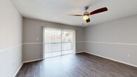 This single-floor residential unit offers a clean, modern aesthetic throughout, characterized by a consistent neutral color palette, wood-look laminate flooring, and textured ceilings. The main living area is bright and open, featuring a ceiling fan with light fixture, a crisp two-tone wall treatment with a dividing chair rail, and large sliding glass doors fitted with vertical blinds that invite natural light and provide direct access to an outdoor space with views of surrounding greenery. The galley-style kitchen is efficiently designed with white cabinetry lining both sides, white appliances including a refrigerator, electric range, and oven, complemented by light wood-look flooring and a single basin sink. Additional rooms echo the same neutral tones and wood-look flooring, each equipped with ceiling fans and two-tone wall finishes, offering versatile, well-lit spaces. The bathroom features a granite-look countertop vanity with an undermount sink, a large mirror, louvered cabinet doors, and textured white walls, creating a clean and functional space. Overall, the unit presents a cohesive, move-in-ready interior with practical finishes and comfortable natural light throughout.