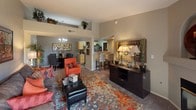 This single-floor residence showcases an open-concept layout that seamlessly connects the living, dining, and kitchen areas into one cohesive and functional space. The living area is anchored by plush carpeting layered with a bold, multicolored area rug featuring warm orange and gray tones that complement the neutral gray walls throughout. A mix of seating, including a vibrant orange accent chair and a tufted leather ottoman, creates an inviting and visually dynamic arrangement. A dark wood credenza with decorative lamps and a framed mirror adds an elegant focal point along one wall.

The dining area, positioned adjacent to the living space, features a round table with upholstered chairs beneath a classic chandelier, enhancing the refined character of the room. Elevated shelving above the main living area provides both decorative and practical storage while adding verticality to the space.

The kitchen is partially open to the main living area via a raised breakfast bar with bar seating, offering a casual dining option. Light wood cabinetry and visible appliances suggest a functional, well-equipped kitchen. The overall ambiance is warm, cohesive, and thoughtfully designed, balancing comfort with a modern aesthetic.