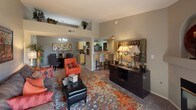 This single-floor residence showcases an open-concept layout that seamlessly connects the living, dining, and kitchen areas into one cohesive and functional space. The living area is anchored by plush carpeting layered with a bold, multicolored area rug featuring warm orange and gray tones that complement the neutral gray walls throughout. A mix of seating, including a vibrant orange accent chair and a tufted leather ottoman, creates an inviting and visually dynamic arrangement. A dark wood credenza with decorative lamps and a framed mirror adds an elegant focal point along one wall.

The dining area, positioned adjacent to the living space, features a round table with upholstered chairs beneath a classic chandelier, enhancing the refined character of the room. Elevated shelving above the main living area provides both decorative and practical storage while adding verticality to the space.

The kitchen is partially open to the main living area via a raised breakfast bar with bar seating, offering a casual dining option. Light wood cabinetry and visible appliances suggest a functional, well-equipped kitchen. The overall ambiance is warm, cohesive, and thoughtfully designed, balancing comfort with a modern aesthetic.