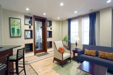 Located at 3100 N William Styron Square in Newport News, Virginia, this single-floor community clubhouse offers a thoughtfully designed gathering space that blends comfort and functionality. The main lounge area features warm hardwood flooring, recessed lighting, and an elegant pendant light fixture that anchors the room. Plush, richly upholstered seating — including classic armchairs and a sofa arranged around a coffee table — creates an inviting atmosphere for relaxation and socializing. Large windows dressed with dark curtain panels allow ample natural light to fill the space while lending a refined aesthetic. A round wooden dining table with carved chairs provides an additional seating area, ideal for casual gatherings. Adjacent to the lounge, a refreshment and service counter is fitted with dark granite countertops, warm wood cabinetry, a toaster oven, microwave, and a coffee maker, making it well-suited for community breakfasts or events. A wall-mounted flat-screen television with a soundbar adds a modern entertainment element. Open shelving stocked with snacks and supplies rounds out the functional amenities. The overall ambiance is warm, polished, and community-focused, offering residents a welcoming retreat for both quiet moments and social interaction.
