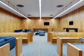 Located at 8 S Sixth St in Council Bluffs, Iowa, this single-floor mock courtroom offers a generously sized, professionally designed space ideal for legal education, training, and simulation. The room is fully enveloped in warm, honey-toned wood panel walls arranged in a geometric relief pattern, creating a formal yet refined architectural atmosphere throughout.

The main courtroom area is anchored by a raised judge's bench at the front, flanked by a witness stand and clerk's station, all finished in matching wood millwork. A wall-mounted flat-screen display is positioned prominently behind the bench, supporting modern presentations and evidence display. Opposing counsel tables are positioned centrally, with a dedicated podium/lectern featuring built-in storage for presenters.

A raised jury box along one side accommodates jurors, outfitted with blue upholstered seating. The gallery features traditional wood pew-style benches for observers. Flexible lounge zones throughout the space include clusters of contemporary blue upholstered chairs and sofas, along with casual seating arrangements for breakout use. A pool table and additional lounge furnishings are present in the rear area of the space.

Overhead, linear LED lighting runs the length of the ceiling, providing consistent, bright illumination across the entire floor. Blue-toned carpeting ties the space together, complementing the wood tones and blue furnishings for a cohesive, professional aesthetic.