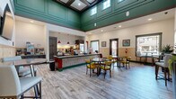 Located at Silver Collection in Fredericksburg, Virginia, this single-floor community dining and lounge space delivers a welcoming, café-style atmosphere with a thoughtfully designed interior. The room is immediately distinguished by its soaring coffered ceiling, finished in a rich teal green with clerestory windows that flood the space with natural light from above. A bold color palette of teal, red, and green carries throughout, from the accent trim to the centrally positioned buffet counter — a tiled, red-based service island equipped with warming vessels and preparation areas, illuminated by striking red pendant lights above. Light wood-plank flooring runs throughout, lending warmth and cohesion to the open layout. Seating options are varied and well-distributed, including round dining tables paired with vibrant yellow upholstered chairs, as well as bistro-height tables positioned near windows with views of the surrounding community. Natural wood wainscoting lines the lower walls, complementing the light and airy feel of the upper walls adorned with colorful framed artwork. A bold "Silver Collection" branded entry mat greets visitors at the red-framed glass entry door, which opens to an outdoor balcony area. Wall-mounted flat-screen televisions enhance the functionality of this space as both a dining and social gathering venue.