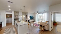 This well-designed single-floor residence offers a thoughtfully planned open-concept layout that seamlessly connects the kitchen, dining, and living areas. The kitchen is a functional centerpiece, featuring stainless steel appliances, white cabinetry, a granite-topped island with a built-in sink and bar seating, and stylish pendant lighting overhead. Light wood-style flooring flows consistently throughout the space, enhancing the bright, cohesive aesthetic.

The living area is generously proportioned, bathed in natural light from large sliding glass doors that open onto a private balcony. The balcony provides outdoor seating and open views of the surrounding neighborhood. A ceiling fan is installed in the living room, adding both practicality and a casual charm.

The residence includes two bedrooms, each featuring walk-in closets, ceiling fans or light fixtures, and windows dressed with blinds and curtains that allow for adjustable natural light. The bedrooms offer a calm, neutral palette with warm wood flooring underfoot.

A dedicated in-unit laundry area with a full-size washer and dryer is conveniently accessible from the kitchen. Built-in shelving near the laundry space provides additional storage. Throughout the residence, neutral wall tones, recessed lighting, and clean architectural lines contribute to a modern yet comfortable living environment.