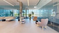 Located at 1250 Avenue Thérèse-Lavoie-Roux in Montréal, this contemporary co-working space offers a thoughtfully designed, open-plan environment ideal for collaboration and productivity. The single-floor layout is generous in scale, providing ample room for a variety of work and lounge configurations.

Architecturally, the space is defined by its striking floor-to-ceiling glass curtain walls that flood the interior with natural light and offer expansive views of the landscaped courtyard and surrounding urban environment. Exposed concrete columns punctuate the open floor plan, adding an industrial yet refined character that contrasts beautifully with the clean, light-toned polished floors.

The space is divided into distinct functional zones. A lounge area features modern upholstered seating — including curved armchairs and a modular sofa — arranged around low accent tables, creating an inviting setting for informal meetings or relaxed work sessions. A dedicated collaborative zone offers light wood tables paired with contemporary olive-green chairs, suitable for group work or focused tasks. Open shelving units display reading materials, adding a curated, professional touch.

Interior glass partitions define additional areas within the space while maintaining visual openness and transparency throughout. Lush potted plants are strategically placed to introduce greenery and warmth. Overhead linear LED lighting complements the abundant natural light, ensuring a well-lit atmosphere at all times. The overall ambiance is bright, modern, and welcoming.