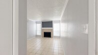 Located at 9655 Chimney Hill Ln in Dallas, Texas, this single-floor unit offers a well-organized layout with distinct living and sleeping areas. The living room serves as the centerpiece of the space, featuring light-toned ceramic tile flooring, crown molding, and a textured ceiling that adds architectural character. A tile-surround fireplace anchors one wall, flanked on both sides by windows dressed with blinds that allow ample natural light to fill the room. The neutral gray walls provide a clean, versatile backdrop throughout. An additional tiled room, accented with a bold gray feature wall, a ceiling fan, and vertical blinds leading to an outdoor-facing door, offers flexible use as a dining area or secondary living space. The bedroom is finished with plush wall-to-wall carpeting and includes a ceiling fan with light fixture and a window for natural light. A generously sized walk-in closet with built-in shelving and hanging rods provides substantial storage. The bathroom is functional and well-equipped, featuring tile flooring, a wide vanity with granite-style countertops and white cabinetry, and a large mirror above. Overall, the unit presents a clean, neutral aesthetic suited for comfortable everyday living.