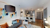 Located at 3310 Woman's Club Dr in Raleigh, NC, this single-floor unit showcases a thoughtfully designed open-concept layout that connects the living, dining, and kitchen areas into one cohesive space. The living area features warm hardwood-style flooring, a bold statement pendant light, and a comfortable seating arrangement anchored by a patterned area rug, creating an inviting atmosphere for relaxing or entertaining.

The kitchen is a standout feature, outfitted with gray cabinetry, light stone countertops, and stainless steel appliances, including a refrigerator and microwave. A peninsula with bar seating provides both additional prep space and a casual dining option, illuminated by sleek pendant lighting overhead. A track lighting fixture adds further brightness to the workspace.

The transition between rooms is smooth and unobstructed, enhancing the sense of openness throughout. Visible from the main living space, an in-unit laundry area with stacked washer and dryer adds everyday convenience. The neutral color palette, accented by vibrant artwork and colorful throw pillows, gives the space a contemporary yet welcoming character. A private entry door completes the layout.