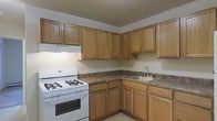 This single-floor space is designed with practicality and functionality in mind. The kitchen serves as a central feature of the home, showcasing warm honey-toned oak cabinetry that lines both the upper and lower walls in an L-shaped configuration, providing generous storage throughout. The dark laminate countertops offer a contrasting look while delivering a durable workspace for meal preparation. A white gas range with a built-in overhead ventilation hood anchors one side of the kitchen, while a stainless steel single-basin sink with a chrome faucet is conveniently positioned along the adjacent wall. The under-cabinet space is fully utilized with matching lower cabinetry, maximizing storage and accessibility. The light-colored flooring and neutral wall tones keep the kitchen feeling open and clean, complementing the warm wood tones of the cabinetry. The overall layout is efficient and well-organized, making it a highly functional cooking and preparation area. The straightforward design of this space prioritizes usability, making it well-suited for everyday residential living.