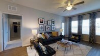 This single-floor, two-bedroom, two-bathroom residence offers a well-balanced layout designed for comfortable everyday living. The living area serves as the heart of the home, featuring warm hardwood-style flooring, neutral gray walls, and a ceiling fan that adds both style and functionality. A gallery wall arrangement above the seating area adds a curated, modern aesthetic, while a large window dressed with patterned curtains invites natural light and creates an airy atmosphere. The living space is generously proportioned, allowing for a full seating arrangement that includes a sofa, accent chairs, and a central coffee table setup. The overall design aesthetic blends contemporary and eclectic elements, with bold accent colors, textured area rugs, and layered lighting that contribute to a warm and inviting ambiance. The neutral color palette throughout provides a versatile backdrop suited to a variety of décor styles. With two full bathrooms and two bedrooms, the floor plan offers both privacy and practicality, making it well-suited for a range of living arrangements. The combination of natural light, thoughtful design details, and a cohesive interior style makes this a comfortable and visually appealing residence.