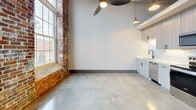This 480 square foot space in Pendleton, SC, showcases a perfect blend of industrial charm and modern functionality. The single-floor layout maximizes efficiency, featuring a hallway and a bathroom. The space is characterized by its loft-like ambiance, with high ceilings adorned with exposed wooden beams and industrial-style pendant lighting.

Large windows flood the area with natural light, offering views of the surrounding landscape. The exposed brick walls add a rustic touch, contrasting beautifully with the smooth, polished concrete floors. The kitchen area boasts sleek, white cabinetry and stainless steel appliances, including a refrigerator and built-in microwave.

The bathroom continues the modern aesthetic with clean lines, a white toilet, and what appears to be marble-like tiling. A ceiling fan enhances air circulation, while the open floor plan allows for versatile use of the space. The industrial-chic design is further emphasized by visible ductwork and electrical conduits, creating a stylish, contemporary atmosphere perfect for those who appreciate minimalist, functional living spaces.