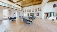 Located at 3389 Endeavor Ln in Vestavia Hills, Alabama, this expansive single-floor community space offers an impressive blend of modern design and warm, inviting aesthetics. The layout is generously proportioned, with light hardwood flooring flowing seamlessly throughout every area.

The living room serves as a dramatic centerpiece, boasting soaring double-height ceilings, an exposed wood-beamed ceiling canopy, and walls of floor-to-ceiling gridded windows that bathe the interior in abundant natural light while framing outdoor views of the surrounding community. Statement pendant lighting in deep navy tones anchors the space, complementing the curated art installations displayed on the crisp white walls.

A large communal kitchen and dining area features an oversized dark-topped island with ample bar seating, warm wood cabinetry, and a striking tiled backsplash. Pendant lights hang above the workspace, and large framed artwork adorns the upper walls. A charming built-in banquette nook with shiplap paneling provides an intimate dining option within the otherwise open layout.

An upper-level mezzanine walkway overlooks the main floor, adding architectural depth. Multiple seating vignettes throughout invite gathering and conversation. The overall ambiance is refined yet welcoming, balancing contemporary finishes with classic architectural details.