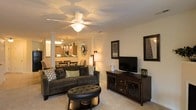 This single-floor space showcases a thoughtfully designed open-concept layout that seamlessly connects the living, dining, and kitchen areas. The living room is anchored by plush, dark upholstered seating, a tufted leather ottoman, and a dark wood media console, creating a warm and inviting atmosphere. A ceiling fan with an integrated light fixture provides both comfort and ambient lighting overhead. Decorative framed artwork and accent pieces add personality to the neutral-toned walls. Soft, light-colored carpeting runs throughout, contributing to the overall warmth of the space. Elegant decorative columns subtly define the transition between the living and dining areas, adding an architectural touch. The dining area, visible from the living room, features a chandelier that lends a refined ambiance to the space. Beyond, the kitchen is equipped with cabinetry and overhead lighting, including a charming chandelier that echoes the home's cohesive design aesthetic. The open sightlines between all areas make the layout feel expansive and well-connected, ideal for both everyday living and entertaining. The overall ambiance is cozy yet polished, with layered lighting and carefully curated furnishings enhancing the inviting character of the space.