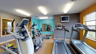 This single-level fitness center is a well-equipped workout space designed to meet essential cardio and strength training needs. The room features a selection of cardio machines, including treadmills and elliptical trainers, arranged to maximize the available floor space while maintaining comfortable circulation between equipment. A wall-mounted flat-screen television provides entertainment for users during workouts.

The interior design incorporates a bold teal accent wall paired with gray-toned walls, creating an energetic yet modern atmosphere conducive to exercise. A large mirror installed along one wall visually expands the space and allows users to monitor their form during workouts, while also reflecting the full range of available equipment. The flooring consists of a durable, low-pile carpeting suitable for a fitness environment.

Overhead, flush-mounted fluorescent light panels provide consistent, even illumination throughout the room. A weight bench and additional strength training equipment are visible near the mirrored wall, offering users versatility in their workout routines. The space is clean, functional, and efficiently organized to serve as a practical fitness facility.