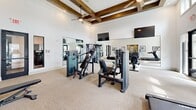 Located at 1540 Edgefield Dr in Garland, TX, this fitness center offers a thoughtfully designed exercise room that balances functionality with attractive architectural character. The space is defined by soaring ceilings adorned with a distinctive coffered framework of rich, dark wood beams, lending a refined aesthetic to what is otherwise a clean, modern interior. Bright white walls and neutral carpeting keep the atmosphere open and energizing.

Large windows positioned at an elevated height along multiple walls invite abundant natural light while providing views of the surrounding outdoor community, creating an uplifting workout environment. Glass panel doors offer direct access to the exterior, further enhancing the connection to the outdoors.

The exercise room is comprehensively equipped with a variety of cardio machines—including treadmills and ellipticals—as well as multiple strength-training stations and cable machines to support full-body workouts. Floor-to-ceiling mirrors line key walls, visually expanding the space and allowing users to monitor their form. Wall-mounted flat-screen televisions add convenience for longer workout sessions. A ceiling fan contributes to comfort, while a built-in storage nook provides organizational utility. Framed athletic artwork on the walls completes the motivating, gym-focused ambiance.