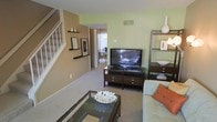 This multi-level home spans three floors, offering a thoughtfully designed layout with distinct living zones connected by a central staircase. The main living area is warm and inviting, featuring soft carpeted flooring, light sage-green accent walls, and neutral warm-toned walls that create a comfortable, cohesive atmosphere. Built-in wall shelving adds both functional storage and a decorative touch, while the open sightline into the dining area enhances the sense of flow and connectivity throughout the space. Natural light filters in from the adjoining dining area, where a large window brightens the room and creates an airy feel. The living room comfortably accommodates a full seating arrangement alongside an entertainment center, making it well-suited for everyday living. The staircase, positioned along the side of the living space, features white railings and leads to the upper and lower floors, providing clear vertical circulation throughout the home. The overall design balances practicality with a casual, welcoming aesthetic, using a palette of earthy tones and soft greens that lend a calm, residential character to the interior. The multi-floor layout offers ample space for a variety of uses across each level.
