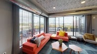This contemporary rooftop amenity space is designed to impress, blending industrial chic aesthetics with comfortable, functional interiors. The lounge area is anchored by a bold coral sectional sofa accented with vibrant yellow and patterned throw pillows, set atop a multi-toned slate tile floor. Distinctive textured tin-style ceiling tiles and exposed industrial ductwork run throughout, lending an urban, loft-inspired character to the entire space.

Floor-to-ceiling glass walls and sliding doors flood the lounge with natural light while opening seamlessly onto a spacious outdoor terrace. The terrace offers sweeping city skyline views, making it an ideal setting for relaxation or social gatherings.

A chevron-patterned accent wall serves as a dramatic focal point in the lounge, featuring a sleek linear electric fireplace and a wall-mounted television, framed by track lighting and a decorative round mirror.

The adjacent dining and kitchen area is equally refined, showcasing a herringbone-patterned hardwood floor, a large communal dining table surrounded by modern navy chairs, and a pendant-lit wall-mounted TV. The fully equipped kitchen includes stainless steel appliances, a refrigerator, microwave, and sink, making the space well-suited for entertaining and events. Together, these areas create a cohesive, upscale communal environment with a vibrant and welcoming ambiance.