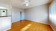 Located at 3734 W El Segundo Blvd in Hawthorne, California, this single-floor unit offers an efficient and functional layout encompassing a living room, kitchen, bathroom, and closet. The living room features durable wood-style laminate flooring in a warm gray tone, neutral painted walls with clean white baseboards, a ceiling fan with a light fixture, and a large window dressed with vertical blinds that allows natural light to fill the space. A warm-toned wood entry door adds a welcoming contrast to the room's neutral palette. A partial wall divides the living area, behind which a wall-mounted HVAC unit and a closet with open shelving provide added utility. The kitchen is thoughtfully appointed with white cabinetry — both upper and lower — granite-patterned countertops, a stainless steel sink, and a white gas range with four burners. White tile flooring grounds the kitchen space cleanly. The bathroom maintains a bright, all-white aesthetic with tile flooring, a combination tub and shower with a handheld showerhead, a white vanity with storage, a wall mirror, and a small window above the tub that introduces natural light. The overall design is clean, practical, and well-maintained throughout.