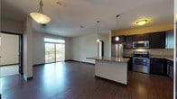 This single-floor residence at 20700 Swenson Dr in Waukesha, Wisconsin offers a well-designed open-concept layout that seamlessly connects the living and kitchen areas. Rich, dark hardwood-style flooring runs throughout the main living spaces, providing a warm contrast to the neutral-toned walls and creating a cohesive, modern aesthetic.

The kitchen is a standout feature, equipped with a full suite of stainless steel appliances including a refrigerator, range, microwave, and dishwasher. Dark espresso cabinetry pairs with granite countertops, and a central island with pendant lighting above offers additional prep space and casual seating. In-unit laundry is conveniently accessible off the kitchen area.

The living area is generously proportioned, filled with natural light from a large sliding glass door that opens to a private balcony with outdoor views. The bedroom features carpeted flooring and a well-sized window with blinds. Two bathrooms are present; one includes a shower-tub combo and dark-framed vanity mirror, while the other offers a spacious layout with a sliding glass shower enclosure and a wide granite-topped vanity. The overall ambiance is clean, contemporary, and functional.