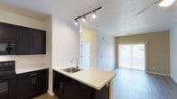 Located at 9444 East 108th Avenue in Henderson, Colorado, this single-floor residence offers a thoughtfully designed open-concept layout that seamlessly connects the kitchen and living areas. The kitchen is a functional centerpiece, anchored by a peninsula-style island with a light countertop, undermount sink, and integrated dishwasher. Dark cabinetry provides a striking contrast against the neutral walls and light-toned flooring, while a sleek track lighting system overhead ensures excellent task illumination. The open flow from the kitchen draws the eye into the spacious living area, which benefits from ample natural light streaming through a large sliding glass door fitted with vertical blinds — offering both privacy and easy access to the outdoors. The neutral color palette throughout creates a clean, versatile backdrop, while the smooth transition between spaces gives the home an airy, connected feel. Light wood-style flooring runs through the main living area, complementing the overall modern aesthetic. A doorway visible from the main living space hints at additional rooms beyond, rounding out this well-organized and inviting single-level home.