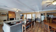 This single-floor space combines rustic charm with a welcoming, lived-in character. The main living area is anchored by warm wood-paneled walls and classic terracotta tile flooring that extends throughout, giving the space a warm, earthy tone. A brick-surround fireplace serves as a focal point on one wall, accompanied by a wall-mounted flat-screen television, making it both functional and inviting.

The ceiling features exposed beams and a textured finish, adding architectural interest overhead, while a ceiling fan ensures comfortable air circulation. Natural light flows generously into the room through a set of white-framed French doors, creating a bright and airy atmosphere despite the warm, wood-toned interior palette.

A long wooden console table is centrally positioned behind the seating area, styled with decorative lanterns and accessories that add to the cozy ambiance. The living space is furnished with a sectional sofa and complementary seating arranged for comfortable gathering. A small dresser and a full-size refrigerator are also visible, suggesting a versatile, multi-use layout.

Overall, the space presents a rustic yet functional character, blending traditional materials with practical amenities.
