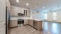 This contemporary single-floor residence offers a thoughtfully designed open-concept layout that seamlessly connects the kitchen and living areas. The kitchen is a standout feature, showcasing light wood-toned cabinetry with glass-front upper panels, a full suite of stainless steel appliances including a range, built-in microwave, refrigerator, and dishwasher, and a peninsula with a light stone countertop and undermount sink. A pair of brushed brass pendant lights add a warm, modern accent above the peninsula. Wood-look plank flooring runs throughout the main living areas, lending a clean and cohesive aesthetic.

The living area is generously proportioned, bathed in natural light from large windows fitted with blinds, and includes a ceiling fan for added comfort. Bedrooms continue the warm palette with plush wall-to-wall carpeting and ceiling fans, creating a cozy retreat. One bedroom features access to a spacious walk-in closet outfitted with built-in shelving and hanging rods along multiple walls.

The bathroom is well-appointed with a dual-sink vanity topped with stone counters, light wood cabinetry, a large mirror, and a combination tub and shower with tile surround. Recessed lighting throughout enhances the bright, airy ambiance of the entire space.