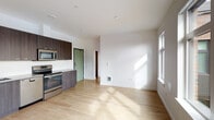 Located at 8220 SE 6th Ave in Portland, Oregon, this single-floor residence offers a clean, contemporary design with consistent light wood flooring and a crisp white color palette throughout. The open-concept living and kitchen area forms the heart of the unit, where a well-appointed kitchen stands out with dark wood-grain cabinetry extending to the ceiling, a white subway tile backsplash, and a full suite of stainless steel appliances including a refrigerator, electric range, built-in microwave, and dishwasher. Generous counter space and ample storage make the kitchen both functional and stylish. The living area flows naturally from the kitchen, benefiting from natural light streaming through a large window along one wall, creating a bright and welcoming atmosphere. The bedroom is a standout space, featuring an expansive picture window that frames views of the surrounding Portland neighborhood, filling the room with abundant daylight. The bathroom is finished with matching dark wood-grain cabinetry, a single vanity with an undermount sink, a large mirror with integrated lighting, and a full tub-shower combination. Recessed lighting throughout the unit maintains a modern, uncluttered aesthetic, and the consistent flooring ties each space together seamlessly.