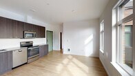 Located at 8220 SE 6th Ave in Portland, Oregon, this single-floor residence offers a clean, contemporary design with consistent light wood flooring and a crisp white color palette throughout. The open-concept living and kitchen area forms the heart of the unit, where a well-appointed kitchen stands out with dark wood-grain cabinetry extending to the ceiling, a white subway tile backsplash, and a full suite of stainless steel appliances including a refrigerator, electric range, built-in microwave, and dishwasher. Generous counter space and ample storage make the kitchen both functional and stylish. The living area flows naturally from the kitchen, benefiting from natural light streaming through a large window along one wall, creating a bright and welcoming atmosphere. The bedroom is a standout space, featuring an expansive picture window that frames views of the surrounding Portland neighborhood, filling the room with abundant daylight. The bathroom is finished with matching dark wood-grain cabinetry, a single vanity with an undermount sink, a large mirror with integrated lighting, and a full tub-shower combination. Recessed lighting throughout the unit maintains a modern, uncluttered aesthetic, and the consistent flooring ties each space together seamlessly.