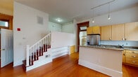 This single-floor residence combines classic architectural character with functional modern updates throughout its thoughtfully arranged layout. Rich hardwood flooring flows consistently through the main living areas, reflecting natural light and adding warmth to the overall ambiance.

The open-concept kitchen is a standout feature, equipped with light maple cabinetry, granite countertops, stainless steel appliances including a French door refrigerator and dishwasher, and a peninsula that provides additional prep space and a natural divide between the kitchen and living area. Tall, multi-pane windows flood the kitchen with abundant natural light, creating a bright and airy atmosphere.

The main living area is spacious and versatile, featuring track lighting along the ceiling, a ceiling fan for year-round comfort, and a window that frames an exterior view. A classic staircase with white-painted balusters and rich wood handrails adds architectural interest and leads to additional lower-level space.

The entry area features a traditional paneled door with a transom window above, lending charm and character to the home's welcome. A compact bathroom completes the layout, finished with tile flooring, a granite-topped vanity with an oval mirror, open wooden shelving for storage, and coordinating wood-trimmed doors that echo the home's consistent design language.