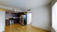 This single-floor space offers a clean, modern aesthetic with an open-concept layout that seamlessly connects the kitchen and living areas. The kitchen is a standout feature, boasting rich dark wood cabinetry that contrasts beautifully against light-toned walls. Stainless steel appliances — including a full-size refrigerator, gas range, and dishwasher — lend a sleek, contemporary feel, while recessed track lighting above the kitchen ensures ample illumination for cooking and entertaining. The countertops are finished in a dark tone that complements the cabinetry, and a functional sink with under-cabinet storage maximizes utility. Light natural hardwood flooring runs throughout the open living space, adding warmth and continuity. A ceiling-mounted HVAC vent and a wall thermostat indicate a centralized climate control system for year-round comfort. The space transitions through a doorway into an adjacent room, suggesting a private bedroom or additional living area beyond. Crisp white walls and smooth ceilings enhance the sense of openness and brightness throughout, creating an inviting and functional environment well-suited for everyday living.