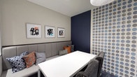 This compact yet thoughtfully designed space offers a versatile environment suited for intimate meetings or casual gatherings. The single-floor layout is anchored by a clean white table positioned at the center, paired with a built-in upholstered banquette along one wall and a studded gray accent chair, creating a balanced seating arrangement.

The design palette centers on a sophisticated combination of navy blue, warm gray, and pops of orange. A deep navy accent wall serves as a striking focal point, complemented by an adjacent wallcovering featuring a bold, graphic dot pattern in tones of blue, gray, and taupe, adding visual depth and texture to the space.

Three framed abstract artworks hang above the banquette, reinforcing the contemporary aesthetic with expressive black, white, and orange brushwork. Coordinating throw pillows in geometric and solid patterns add layers of comfort and style.

Overhead, a modern pendant light fixture provides focused illumination. The overall ambiance is polished and intentional, blending functional furnishings with design-forward elements to create an inviting and visually engaging environment.