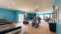 This single-floor fitness center offers a generously sized workout space designed to accommodate a comprehensive range of exercise equipment and training activities. The interior is immediately distinguished by its bold teal accent walls, which contrast with the white ceiling and create an energizing atmosphere conducive to physical activity.

The space is organized into distinct functional zones. The cardio area features multiple treadmills and elliptical machines from the TRUE Fitness brand, arranged to allow comfortable spacing between users. Adjacent to this zone, a dedicated strength training section houses a variety of cable machines, multi-use weight stations, and adjustable benches, all positioned on protective rubber floor mats.

Natural light enters through several windows fitted with white blinds, supplemented by recessed ceiling lighting throughout. Ceiling fans provide additional air circulation across the main workout area. A large wall mirror near the entrance enhances the sense of space while allowing users to monitor their form. The floor is finished with a durable, cork-toned carpeted surface suited for high-traffic fitness use. An exterior-access door provides convenient entry and exit directly from outside.