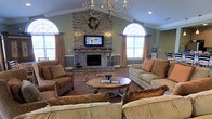 This single-floor space centers around a striking floor-to-ceiling stone fireplace, which serves as the room's focal point and adds a sense of rustic elegance. A wall-mounted television above the fireplace mantel is framed by decorative sconce lighting and seasonal accents, enhancing the hearth's visual impact. The vaulted ceiling rises generously overhead, amplifying the sense of openness and grandeur, while an elegant chandelier anchors the space from above. Two large arched windows flank the fireplace, each dressed with warm, earth-toned drapes that complement the sage green walls and neutral carpeting throughout. The seating arrangement is thoughtfully designed for comfort and conversation, featuring multiple upholstered sofas and armchairs in warm brown tones surrounding a classic wooden oval coffee table. A patterned area rug grounds the central seating area, adding texture and warmth. Crown molding along the upper walls contributes a refined, finished aesthetic. The overall ambiance is cozy yet spacious, blending traditional architectural details with comfortable furnishings to create an inviting communal atmosphere suited for relaxation and social gathering.