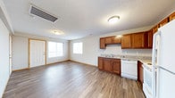 This single-floor space offers a practical and well-organized open-concept layout that seamlessly connects the living area with the kitchen. The flooring throughout is a durable hardwood-style laminate in a warm gray-brown tone, providing a clean and cohesive look across the entire space.

The kitchen is outfitted with rich, warm-toned wooden cabinetry offering ample upper and lower storage. A granite-style countertop runs along the base cabinets, complemented by a double-basin sink and a white dishwasher, making the space both functional and visually inviting.

The living area is open and airy, benefiting from two windows that allow natural light to fill the room. A standard entry door with a sidelight panel provides additional brightness and a welcoming entryway. The neutral beige walls pair well with the wood tones and lighter flooring, creating a balanced, understated aesthetic.

The ceiling features a textured finish with flush-mount light fixtures providing ample overhead illumination. Ventilation is handled by ceiling-integrated HVAC vents, ensuring year-round comfort. Baseboards trim the perimeter of the room neatly, adding a finishing touch to the overall design. The layout is efficient, making excellent use of the available space.