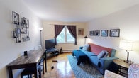 This single-floor space centers around a bright and inviting living room that showcases classic parquet hardwood flooring, lending warmth and character to the interior. A centrally positioned window dressed with rich, dark curtains allows natural light to filter in, illuminating the room's neutral, cream-toned walls. The living area is thoughtfully arranged, featuring a comfortable seating area with a blue sofa and a coral-toned accent piece, complemented by a patterned area rug that anchors the space. A flat-screen television sits on a media stand along one wall, while a tall floor lamp and an additional accent lamp provide flexible lighting options. A row of framed artwork lines the far wall, adding a personalized, curated touch to the décor. A baseboard heating unit runs along the base of the window wall, indicating a practical heating setup. Adjacent to the living area, a dark-toned table and shelving unit provide additional workspace or dining utility. Overall, the space presents a functional and comfortable layout with a lived-in, homey ambiance.