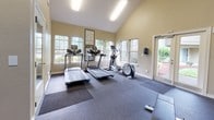 This fitness center offers a thoughtfully designed workout environment that balances functionality with an open, airy atmosphere. The standout architectural feature is the dramatic vaulted ceiling, which lends a spacious, expansive feel to the entire room. Large multi-pane windows line the walls, flooding the space with natural light and offering views of the surrounding greenery, while French doors provide convenient direct access to the outdoors.

The facility is outfitted with a comprehensive selection of equipment to support a full-body workout routine. Cardio options include two treadmills and an elliptical trainer, all positioned to take advantage of the natural light from the windows. A dedicated strength training area features professional Precor functional training machines, a full rack of dumbbells ranging across multiple weight increments, and a flat workout bench for free weight exercises. A large industrial fan provides additional airflow circulation throughout the space.

The flooring consists of durable rubber gym mats layered over a dark base floor, providing the traction and impact absorption needed for a variety of exercises. Wall-mounted flat-screen televisions are positioned for easy viewing during workouts. The warm neutral-toned walls and well-lit environment create an inviting and motivating atmosphere for exercise.