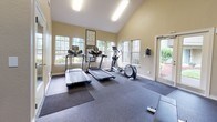 This fitness center offers a thoughtfully designed workout environment that balances functionality with an open, airy atmosphere. The standout architectural feature is the dramatic vaulted ceiling, which lends a spacious, expansive feel to the entire room. Large multi-pane windows line the walls, flooding the space with natural light and offering views of the surrounding greenery, while French doors provide convenient direct access to the outdoors.

The facility is outfitted with a comprehensive selection of equipment to support a full-body workout routine. Cardio options include two treadmills and an elliptical trainer, all positioned to take advantage of the natural light from the windows. A dedicated strength training area features professional Precor functional training machines, a full rack of dumbbells ranging across multiple weight increments, and a flat workout bench for free weight exercises. A large industrial fan provides additional airflow circulation throughout the space.

The flooring consists of durable rubber gym mats layered over a dark base floor, providing the traction and impact absorption needed for a variety of exercises. Wall-mounted flat-screen televisions are positioned for easy viewing during workouts. The warm neutral-toned walls and well-lit environment create an inviting and motivating atmosphere for exercise.