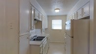 This single-story space includes a galley-style kitchen designed for efficient use of its layout. The kitchen features white cabinetry lining both walls, providing ample storage through upper and lower cabinets with traditional knob hardware. A double-basin sink sits along one wall beneath the upper cabinets, accompanied by a continuous light-colored countertop that extends the length of the kitchen. A full-size refrigerator anchors the opposite wall, complementing the overall white-and-neutral color palette. A range hood is integrated above the cooking area, and a small window at the far end of the kitchen allows natural light to filter in, brightening the space. The floors are a light, neutral tone that blends seamlessly with the cabinetry and walls. A flush-mount ceiling light fixture provides additional illumination, and the textured popcorn ceiling adds a classic finish overhead. Crown molding along the upper edges of the walls adds a subtle architectural detail. The overall ambiance is clean, functional, and understated, with a cohesive neutral palette throughout.