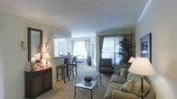 This single-floor residence offers a thoughtfully designed layout that seamlessly blends comfort and functionality. The living area serves as the heart of the space, featuring plush carpeting, crown molding, and smooth white ceilings that contribute to a clean, classic aesthetic. A generous sofa anchors the seating arrangement around a decorative coffee table, while an upholstered armchair adds additional seating near the windows.

Natural light flows freely throughout the space, courtesy of multiple large windows dressed with layered curtains in neutral tones. A sliding glass door provides direct access to an outdoor area, further enhancing the bright and airy atmosphere. Lush indoor greenery complements the natural light, adding a refreshing organic element to the décor.

A breakfast bar with bar-height seating creates a natural transition between the living area and the adjacent dining or kitchen space. Wall sconces and table lamps provide warm ambient lighting throughout. A dark wood dresser with a mirror adds a classic touch near the entryway, rounding out the elegant yet approachable character of this well-maintained single-level residence.