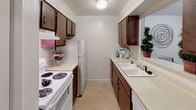 This single-floor unit features two bedrooms, one full bathroom, and a half bathroom, offering a straightforward and practical layout. The living and dining areas are carpeted and connected, with natural light entering through multiple windows fitted with blinds. The dining space is defined by a pass-through opening to the kitchen, creating a convenient connection between the two areas. Each bedroom is carpeted and includes a ceiling fan with a light fixture. One bedroom features a window for natural light and a standard closet with built-in shelving and a hanging rod, while the second bedroom also includes a ceiling fan and access to an additional closet. The galley-style kitchen is equipped with dark wood cabinetry on both sides, a white electric range, refrigerator, and a double-basin sink. Light tile flooring runs throughout the kitchen. The full bathroom features tile flooring, a vanity with countertop space, and a shower-tub combination. The half bathroom provides a toilet and vanity. The unit's neutral white walls and light-toned finishes throughout contribute to a clean, functional living environment.