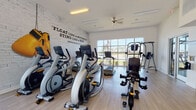 This single-floor fitness center is designed to inspire and accommodate a wide range of workout routines. The main exercise area is outfitted with a comprehensive selection of Matrix-brand cardio equipment, including treadmills, elliptical trainers, and a stair climber, all arranged to maximize floor space and usability. A dedicated strength zone features a multi-station cable machine, adjustable weight bench, and a full rack of dumbbells, providing everything needed for resistance training. Large floor-to-ceiling windows flood the space with natural light while offering views of the surrounding outdoor landscape, including what appears to be a pool area. A striking whitewashed brick accent wall bears a bold Muhammad Ali quote — "Float like a butterfly, sting like a bee" — alongside oversized decorative boxing gloves, lending the space a motivational and athletic character. Light wood-look flooring runs throughout, contributing to a clean, contemporary aesthetic. A glass-paneled garage-style door opens the space toward the outdoor pool area, enhancing airflow and versatility. A ceiling fan aids ventilation, and a mounted TV keeps members entertained during workouts. A small service station near the entrance provides cleaning supplies and a compact refrigerator for member convenience. The facility operates 24 hours and is reserved for residents and their guests.