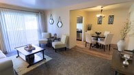 This single-floor residence offers a thoughtfully designed open-concept layout that seamlessly connects the key living areas. The living room is anchored by plush carpeting and features a classic coffee table, accent chairs with decorative upholstery, and a pair of ornamental mirrors adorning the neutral-toned walls. Natural light filters in through a large window dressed with sheer curtains, creating a bright and welcoming atmosphere. Transitioning from the living room, hardwood-style flooring flows into the dining area, where a round wooden dining table surrounded by upholstered chairs serves as an elegant gathering spot beneath a statement chandelier. The open passageway leads directly into the kitchen, which features dark cabinetry, stainless steel appliances, and efficient use of space. The cohesive color palette of warm neutrals and earthy tones throughout the space contributes to a comfortable and inviting ambiance. The open sightlines between rooms foster a sense of connectivity, making the layout both functional and ideal for everyday living and entertaining.