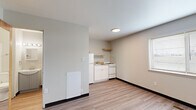Located at 2215 E 56th St in Indianapolis, Indiana, this one-bedroom, one-bathroom unit offers a practical and efficiently designed single-floor layout. Light wood-style flooring runs throughout the space, complementing the neutral, light gray walls and white trim that create a clean, cohesive aesthetic.

The open main area seamlessly integrates the living space with a compact kitchen. The kitchen features white cabinetry, a white refrigerator, a gas range, a single-basin sink, and an open wood shelf above for additional storage. A window fitted with blinds allows natural light to brighten the area.

The spacious bedroom occupies a generous portion of the unit and includes a large window that fills the room with natural light. An open closet with a hanging rod provides accessible storage. Warm wood-toned doors add a touch of contrast to the otherwise light interior.

The bathroom is efficiently appointed with a gray vanity cabinet, a mirrored medicine cabinet with overhead lighting, a toilet, and an additional storage nook. The light wood-style flooring extends into the bathroom, maintaining a consistent look throughout the unit.

A wall-mounted heating unit is present in the main living area, and a fire extinguisher is accessible near the bedroom entry, reflecting attention to safety. The overall design is straightforward, clean, and functional.