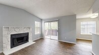 Located at 12801 Roydon Dr in Houston, TX, this single-floor, one-bedroom, two-bathroom home offers a well-considered layout across a compact but efficient floor plan. The living room is a standout space, featuring vaulted ceilings, a striking stone-surround fireplace, and wood-look flooring that runs throughout the main areas. Multiple windows flood the room with natural light, while a sliding glass door opens directly onto the patio, providing easy outdoor access and views of surrounding greenery. The kitchen is equipped with white cabinetry, track lighting, black appliances including a full-size refrigerator, and a breakfast bar that connects to the dining area, creating an open and functional flow. Both bathrooms are finished with wood-look flooring, white vanities with light countertops, framed mirrors, and chrome fixtures. The bedroom features plush carpet, a ceiling fan with light fixture, and a natural light source via a window with blinds. It is complemented by a walk-in closet with wire shelving and hanging rods on multiple levels for organized storage. Additional conveniences include an in-unit laundry area and a private patio. The consistent gray and white palette throughout gives the home a clean, cohesive aesthetic.