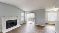 Located at 12801 Roydon Dr in Houston, TX, this single-floor, one-bedroom, two-bathroom home offers a well-considered layout across a compact but efficient floor plan. The living room is a standout space, featuring vaulted ceilings, a striking stone-surround fireplace, and wood-look flooring that runs throughout the main areas. Multiple windows flood the room with natural light, while a sliding glass door opens directly onto the patio, providing easy outdoor access and views of surrounding greenery. The kitchen is equipped with white cabinetry, track lighting, black appliances including a full-size refrigerator, and a breakfast bar that connects to the dining area, creating an open and functional flow. Both bathrooms are finished with wood-look flooring, white vanities with light countertops, framed mirrors, and chrome fixtures. The bedroom features plush carpet, a ceiling fan with light fixture, and a natural light source via a window with blinds. It is complemented by a walk-in closet with wire shelving and hanging rods on multiple levels for organized storage. Additional conveniences include an in-unit laundry area and a private patio. The consistent gray and white palette throughout gives the home a clean, cohesive aesthetic.