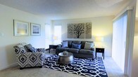 This single-floor space offers a comfortable and thoughtfully designed living environment. The living room serves as the centerpiece, featuring soft, neutral wall tones that create a calm and welcoming atmosphere. Large windows fitted with white horizontal blinds allow generous amounts of natural light to fill the room, enhancing the airy feel of the space.

The living room is furnished with a plush gray sofa accented by decorative pillows, complemented by a patterned armchair in navy and white that adds visual interest. A round metallic coffee table anchors the seating arrangement atop a bold navy and white geometric area rug, which adds a striking contrast against the light carpeted flooring. Two matching dark end tables with warm-toned lamps frame the sofa, providing both symmetry and ambient lighting.

Wall décor includes a triptych artwork depicting stylized bare trees, adding a nature-inspired focal point to the room, alongside a framed landscape print that enhances the curated aesthetic. The textured ceiling adds subtle depth to the overall design.

The space strikes a balance between modern style and everyday comfort, making it a functional and visually appealing environment suited for relaxing or entertaining.